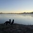 Two black and white dogs standing on a frosty riverbank, looking out over a calm river with morning mist rising and a clear sky at sunrise