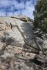 Large textured rock formation surrounded by scattered boulders and dry vegetation under partly cloudy sky with evergreen trees to the right