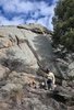 Person sitting on a rocky ledge near a small campfire with a large rock formation and tall pine tree in the background under a partly cloudy sky