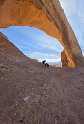 Two dogs walking on a smooth rock surface under a large natural sandstone arch with a blue sky and clouds visible through the arch