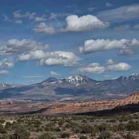Expansive landscape of desert scrub and red rock cliffs with snow-capped mountains under a sky filled with scattered white clouds