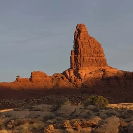 tall reddish rock formation rising from a desert landscape with scattered bushes and a partly cloudy sky