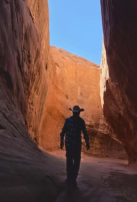 Silhouette of a person wearing a cowboy hat walking through a narrow, sunlit red rock canyon with smooth sandstone walls and a clear blue sky visible above
