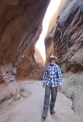 Older man in a plaid shirt and cowboy hat walking through a narrow sandstone canyon with smooth, curved walls