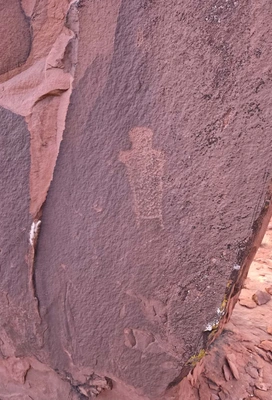 weathered petroglyph carved into a reddish-brown rock surface, showing a simple humanoid figure with outstretched arms