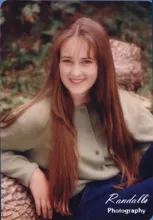 Smiling young woman with long straight hair wearing a light green buttoned sweater sitting outdoors against a log