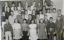 Large group of children and one adult posing together in front of a curtain, dressed in mid-20th century clothing
