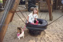 Young girl sitting on a tire swing holding a pink leash attached to a dog wearing a pink bandana in a sandy playground