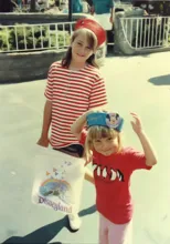 Two children wearing colorful hats and casual clothes, one holding a Disneyland souvenir bag with a castle and rainbow design