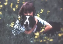Child hugging a black and white dog while lying in a field with yellow flowers