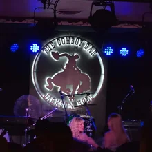 Stage with drum set and microphones in front of a circular illuminated sign showing a cowboy riding a bucking horse with the text The Cowboy Bar Jackson Hole