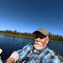 Older man with a white beard wearing a cap and plaid shirt sitting in a boat on a calm lake with a forested shoreline under a clear blue sky