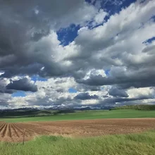 plowed field with green grass and rolling hills under a sky filled with large, dramatic clouds