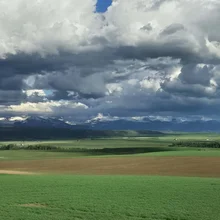 Green and brown fields under a dramatic sky with dark clouds and patches of sunlight highlighting distant mountains