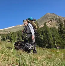 elderly man hiking with a large backpack and walking stick accompanied by a black dog in a grassy mountain area with trees and a peak in the background