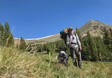 Older man hiking with a large backpack and walking stick accompanied by a black and white dog in a mountainous grassy landscape