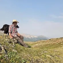 Man in a wide-brimmed hat and plaid shirt sitting on a rocky grassy hill with a black dog beside him, overlooking distant mountains under a clear sky