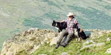 Man in a cowboy hat sitting on a rocky hillside with two black and white dogs resting beside him