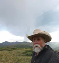 Older man with a white beard wearing a wide-brimmed hat and dark jacket standing in a grassy mountainous landscape under a cloudy sky