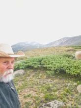 Older man with a white beard wearing a straw hat and dark jacket, standing in a mountainous landscape with green shrubs and rocks