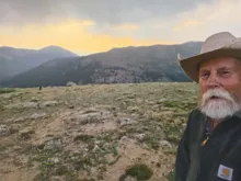 older man with white beard wearing a cowboy hat and a dark jacket standing on rocky mountain terrain at sunset